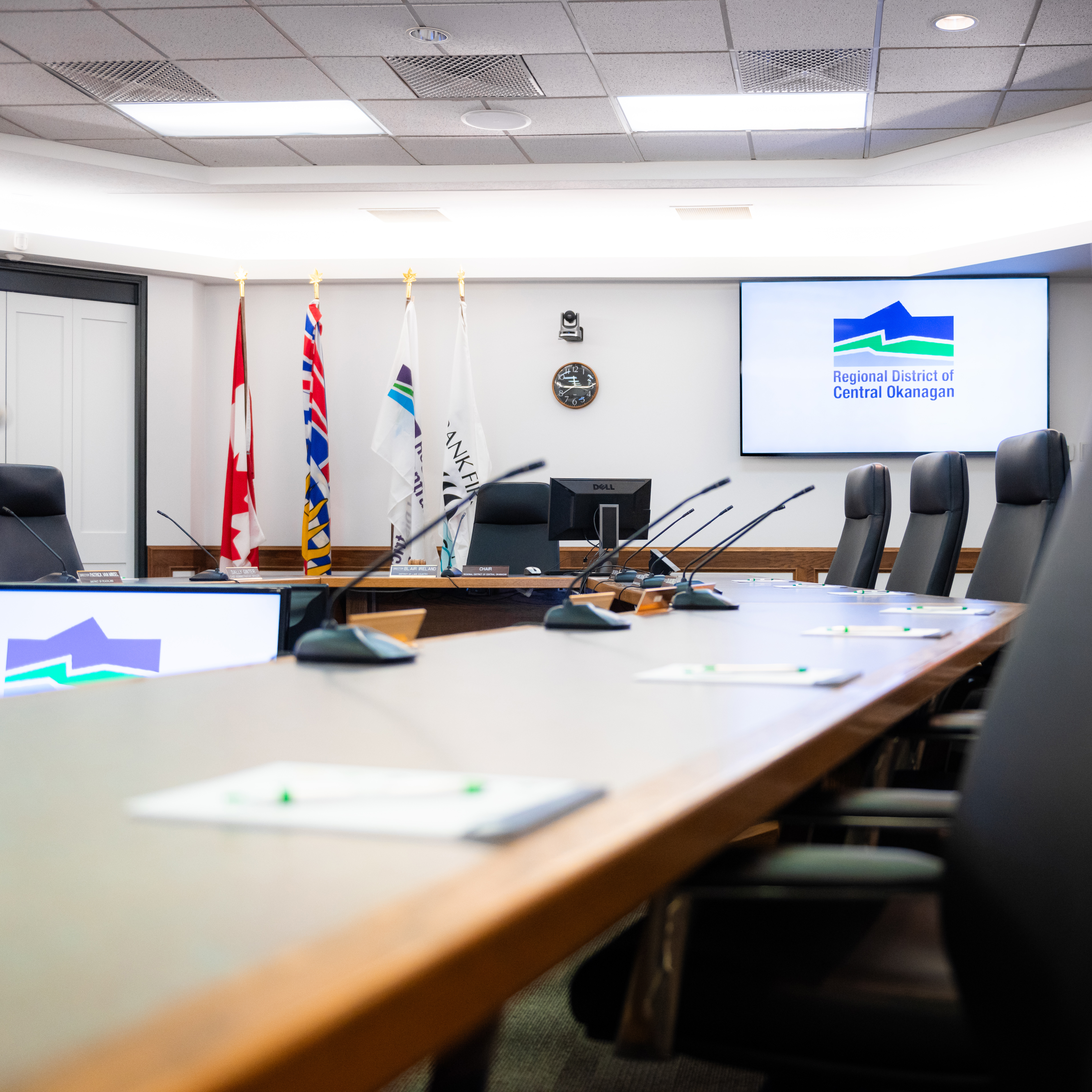 A  boardroom with conference microphones, flags, and a screen displaying the Regional District of Central Okanagan logo