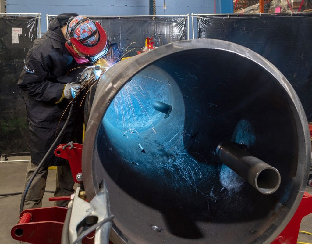 Person welding a large metal pipe with bright sparks inside an industrial workshop.