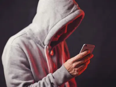 A person wearing a hooded sweatshirt looks down at a smartphone in their hands, partially obscured by shadow against a dark background.