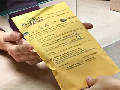 Hands holding a yellow radon test kit instruction sheet labeled ‘Take Action on Radon’ across a counter.